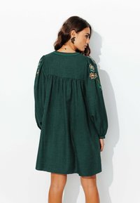 Robe verte à texture avec des manches ballons et des détails floraux brodés sur les épaules. Présente un dos froncé et un col rond.