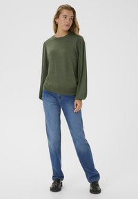 Pull en vert olive avec des manches texturées, col rond et un corps ajusté. Associé à un jean bleu droit et des chaussures noires.