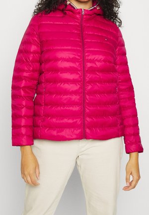 Kobieta ubrana w jasnoróżową pikowaną kurtkę z kapturem, przednim zamkiem błyskawicznym oraz jasnobeżowe spodnie, stojąca na jednolitym tle.