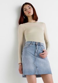 Light beige long-sleeve top paired with a light blue denim mini skirt featuring a raw hem and angled seams. Model stands against a white background.