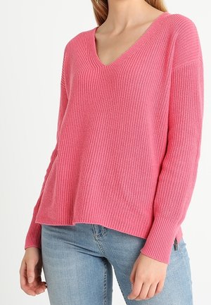 Kvinde iført en lyserød ribstrikket V-hals sweater og lyseblå jeans, vist fra skuldre til hofter mod en hvid baggrund.