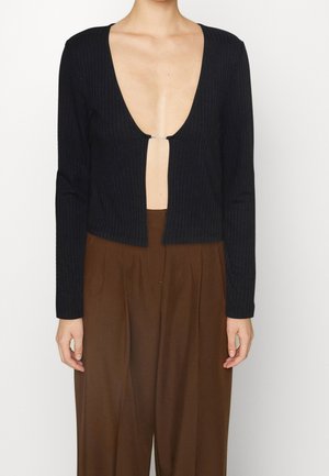 Persona che indossa un cardigan cropped a coste nero con maniche lunghe e scollatura profonda a V, abbinato a pantaloni larghi beige a vita alta.