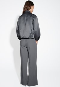 Giacca bomber in satin di colore grigio scuro, con colletto alto, spalle cadenti e vestibilità rilassata, abbinata a pantaloni sartoriali grigi.