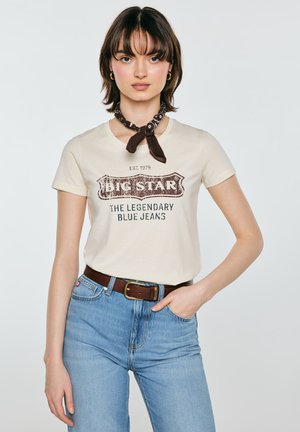 Młoda kobieta w kremowym t-shircie z nadrukiem, niebieskich dżinsach z brązowym paskiem i ciemnobrązowej chustce z wzorem na szyi, stojąca z jedną ręką w kieszeni.