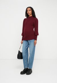 Kvinne i burgunder ribbestrikket genser, lys blå rette jeans, svarte platåsko, holder en svart håndveske, står mot hvit bakgrunn.