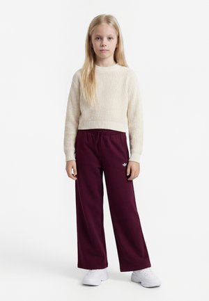 Ung pige med langt blondt hår iført en cremefarvet strikket sweater, burgunderrøde vide bukser og hvide sneakers, stående mod en ensfarvet baggrund.