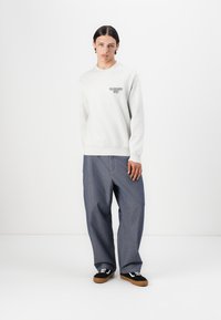 Lys grå sweatshirt med rund hals og trykt tekst på venstre bryst, parret med løse mørkeblå denimbukser og sorte sneakers.