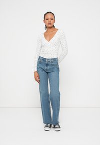 White long-sleeve top with small floral pattern, paired with high-waisted light blue jeans. Model wears black and white sneakers.