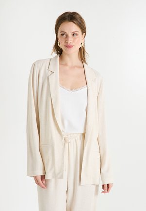 Blazer en lin beige avec une coupe décontractée, des revers crantés et deux poches avant. Porté par-dessus un caraco blanc avec des bordures en dentelle.