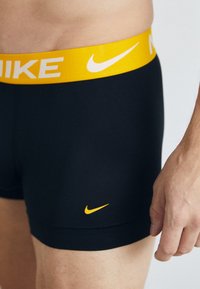 Sorte boxer underbukser med en gul midjebånd som har hvit "NIKE" tekst; liten oransje Nike-logo på venstre side foran.