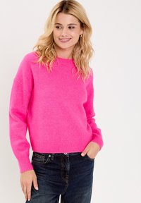 Hellrosa gestrickter Pullover mit rundem Ausschnitt, gerippten Bündchen und Saum. Kombiniert mit dunkelblauen Jeans. Weiche Textur.