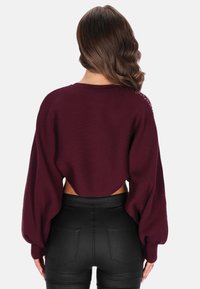 Bordeaux cropped trui met geribbelde textuur, wijde mouwen en open rugdetail, gecombineerd met zwarte aansluitende broek.