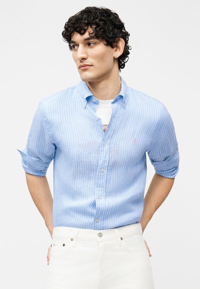 Άνδρας με σγουρά μαλλιά, ντυμένος με ένα ανοιχτό μπλε πουκάμισο με ρίγες πάνω από λευκή μπλούζα και λευκό παντελόνι, κοιτάζει προς τα αριστερά του με τα χέρια πίσω από την πλάτη.