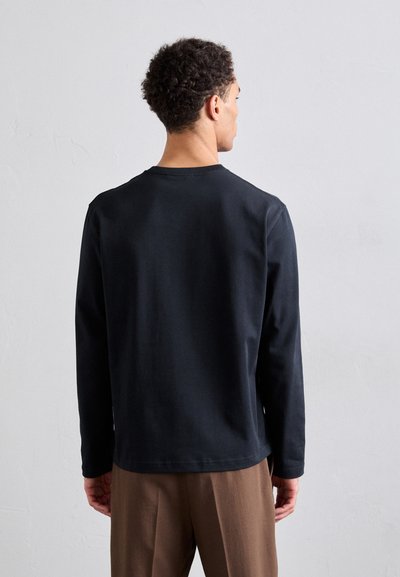 Chemise noire à manches longues en tissu doux avec un col rond. Présente un ourlet droit et des coutures latérales, portée avec un pantalon marron.