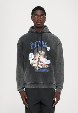 Young man wearing faded black hoodie with colorful rabbit and carrot graphic and text "Family Business" standing against plain background.