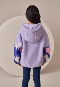 Jachetă din polar lila cu glugă, având modele abstracte colorate pe mâneci. Textură moale, croială supradimensionată și manșete ribbate.