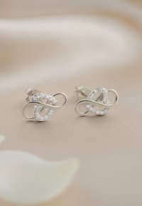 Silberne Ohrstecker mit einem abstrakten, ineinander verflochtenen Design, verziert mit klaren Kristallen, vor einem sanften, neutralen Hintergrund.