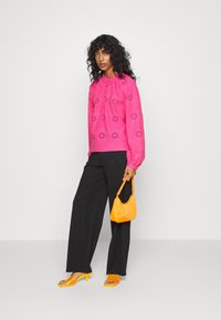 Blusa bordada rosa com mangas bufantes, combinada com calças largas pretas, mala amarela e sapatos de salto aberto amarelos.