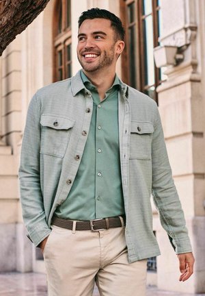 Lächelnder Mann trägt ein hellgrünes Hemd mit Knöpfen, passende Jacke, beige Hose und braunen Gürtel, geht draußen in der Nähe eines Gebäudes.