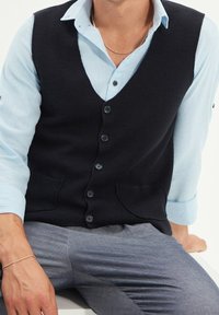 Svart texturerad väst med fem knappar, kombinerad med en ljusblå skjorta med krage. Grå smal passform byxor fulländar outfiten.