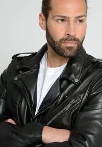 Czarna skórzana kurtka motocyklowa z srebrnymi elementami, kieszeniami na zamek i epoletami na ramionach. Nosi się ją na białej koszuli. Teksturowane wykończenie.