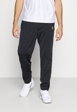 Mann trägt schwarze Sporthose mit Logo am Oberschenkel, weiße Sportschuhe und weißes kurzärmliges Shirt, steht auf einem einfarbigen grauen Boden.