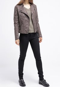 Braune Kunstleder-Motorradjacke mit schrägen Reißverschlüssen, getragen über einem beigefarbenen Stricktop und schwarzen Slim-Fit-Jeans, kombiniert mit schwarzen Schuhen.