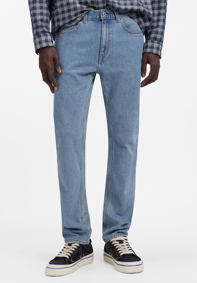 Helerohelised teksapüksid on sirge säärekujundusega, viie taskuga ja pleekinud tekstuuriga. Sobitatud mustade tenniste ja ruudulise särgiga.