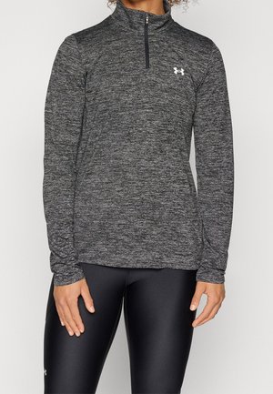 Schwarzes und graues, strukturiertes langärmliges Sportoberteilen mit einem halben Reißverschluss am Kragen und einem Logo auf der oberen linken Brust. Getragen mit schwarzen Leggings.