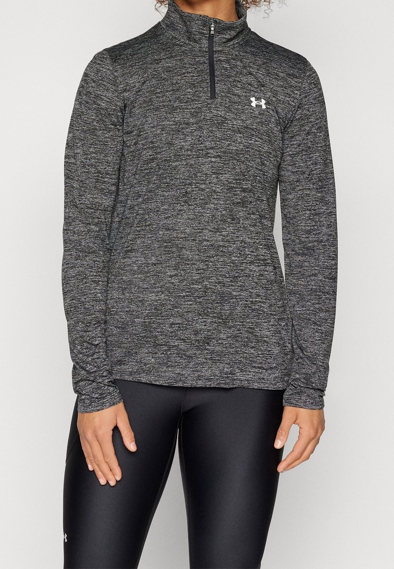 Schwarzes und graues, strukturiertes langärmliges Sportoberteilen mit einem halben Reißverschluss am Kragen und einem Logo auf der oberen linken Brust. Getragen mit schwarzen Leggings.