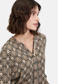Gemusterte Bluse mit einem floralen Design in Schwarz, Creme und Brauntönen. Leichter Stoff, Knopfverschluss und V-Ausschnitt.