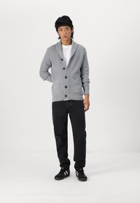 Grå strikket cardigan med sjalkrage, med brune knapper og to frontlommer, kombinert med svarte jeans og svarte sneakers.