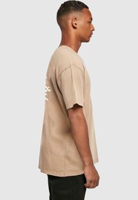 Beige bomulds t-shirt med korte ærmer, løs pasform og tekstdesign på ryggen. Ensartet tekstur uden synlige mønstre eller detaljer.
