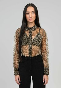 Blouse transparente avec un motif léopard dans les tons brun et beige, dotée d'un col noir avec des boutons décoratifs, associée à un pantalon noir.