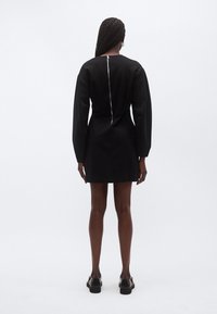 Μαύρο φόρεμα με μακριά, δομημένα μανίκια, εφαρμοστή μέση και φερμουάρ στο πίσω μέρος με περιμετρική ραφή. Το ύφασμα έχει λεία υφή. Μαύρα παπούτσια.