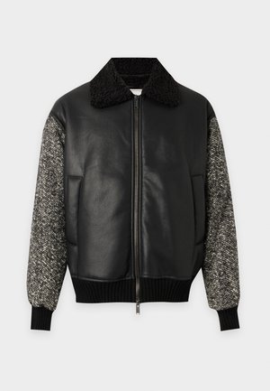 Homme portant une veste bomber en cuir et laine noire, une chemise blanche, un pantalon large marron et des chaussures noires, debout devant un fond blanc.