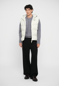 Puffer vest in light beige with a hood, featuring quilted sections, worn over a gray sweater, paired with wide black trousers and dark shoes.