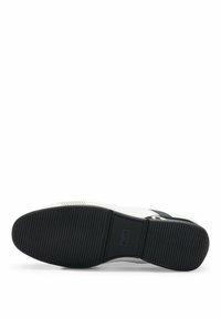 Zapatilla blanca con suela de goma negra. Presenta detalles perforados y un acento a rayas. El talón muestra la marca. Diseño clásico con cordones.