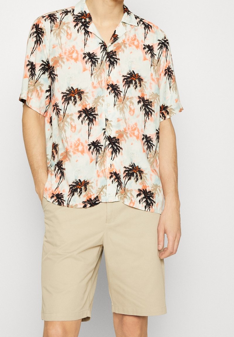 Camicetta a maniche corte con un motivo di palme nei toni del blu chiaro e corallo, realizzata in un tessuto morbido; abbinata a pantaloni bermuda beige con frontale piatto.