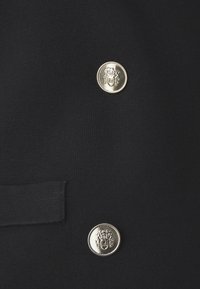 Czarna dwurzędowa marynarka z dwoma ozdobnymi srebrnymi guzikami z herbem oraz boczną kieszenią. Gładki, teksturowany materiał.