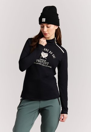Femme portant un bonnet noir, un haut noir à manches longues avec un texte sur le thème du ski, et un pantalon vert, posant devant un fond clair uni.