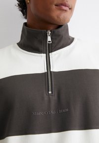 Fehér és sötétszürke negyedcipzáros pulóver, sima anyagból, magas gallérral, valamint a "Marc O'Polo DENIM" felirattal, dombornyomott részletekkel.