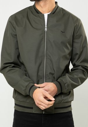 Homme portant une veste bomber vert olive avec des poignets rayés noirs et une ceinture, tenant ses mains jointes devant lui.