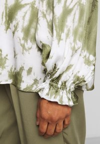 Bluzka z długim rękawem w biało-zielony wzór tie-dye. Materiał jest miękki, z marszczonymi mankietami i luźnym krojem. Zestawiona z oliwkowo-zielonymi spodniami.