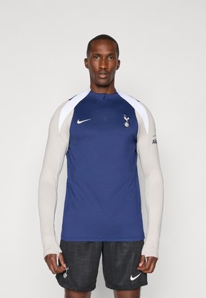 Maillot de sport bleu marine avec un col haut, accents blancs sur les épaules et logo de l'équipe ; associé à un short à carreaux noirs et à une marque Nike.