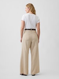 Femme aux cheveux blonds portant un haut blanc à manches courtes rentré dans un pantalon beige taille haute, jambes larges, avec des poches arrière et une ceinture noire.