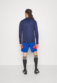 Muž s dredy stojí čelem ke zdi, na sobě má navy modrou dlouhou košili, modré a červené šortky, černé podkolenky a černé kopačky.