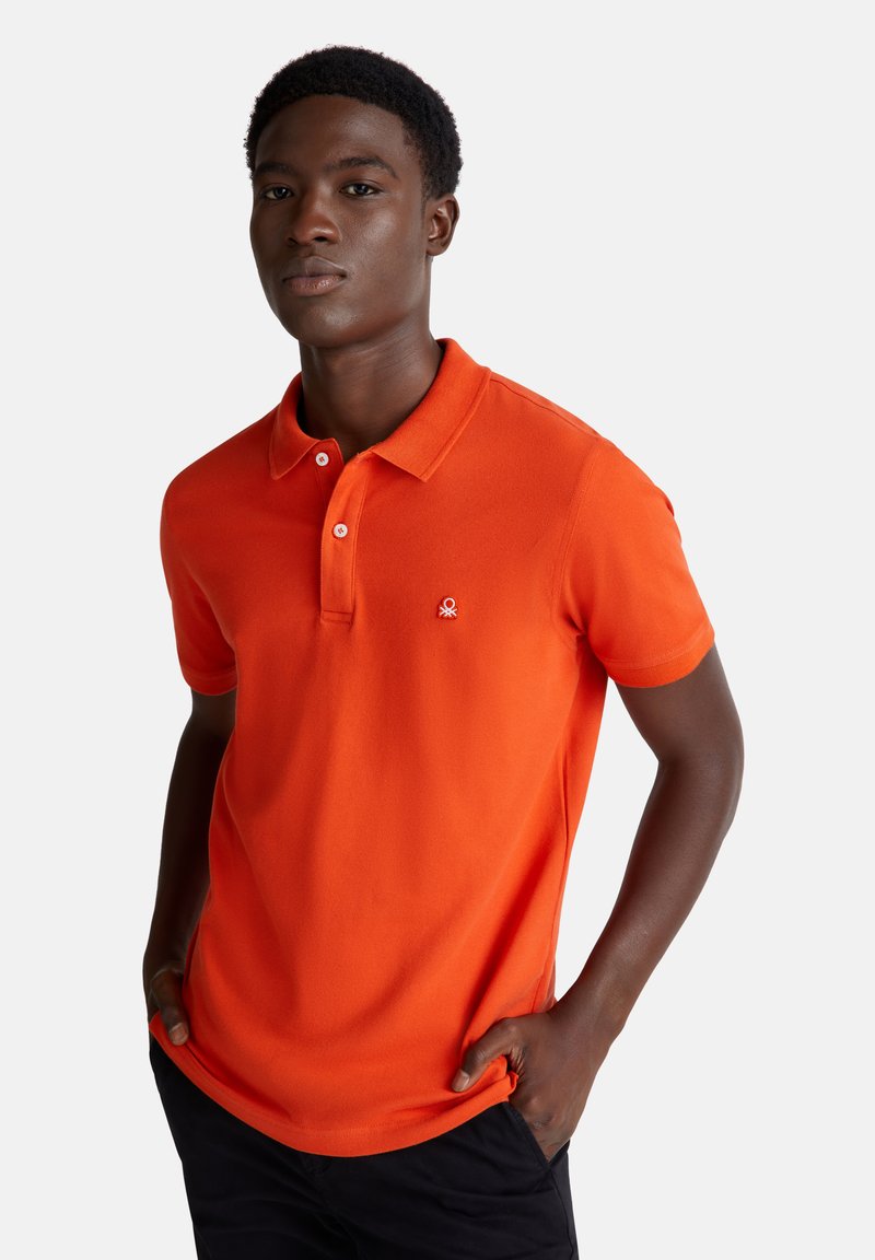 Mladý muž v oranžovém polo tričku s krátkým rukávem a malým logem na hrudi, stojící s rukama v kapsách černých kalhot.
