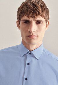 Camicia azzurra chiara con colletto, texture liscia e bottoni blu scuro. Nessun motivo o dettaglio aggiuntivo visibile.