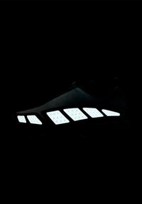 Scarpa atletica nera con cinque forme geometriche bianche riflettenti lungo la suola, esposta su uno sfondo scuro.